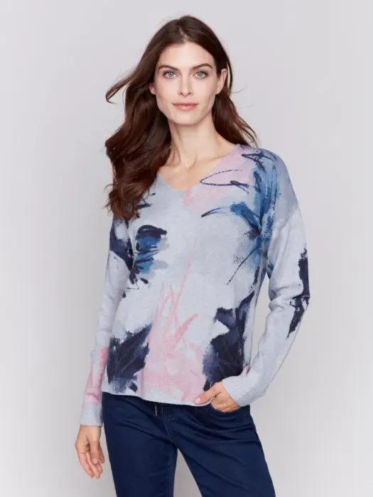 A woman with long brown hair is donning the epitome of winter comfort from Charlie B: a light grey Printed Plushy Sweater featuring an elegant watercolor-style print in pink and blue, perfectly paired with dark blue jeans.