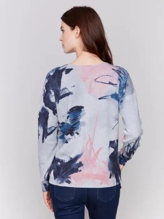 A woman with long brown hair is donning the epitome of winter comfort from Charlie B: a light grey Printed Plushy Sweater featuring an elegant watercolor-style print in pink and blue, perfectly paired with dark blue jeans.