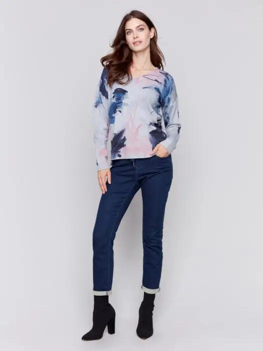 A woman with long brown hair is donning the epitome of winter comfort from Charlie B: a light grey Printed Plushy Sweater featuring an elegant watercolor-style print in pink and blue, perfectly paired with dark blue jeans.