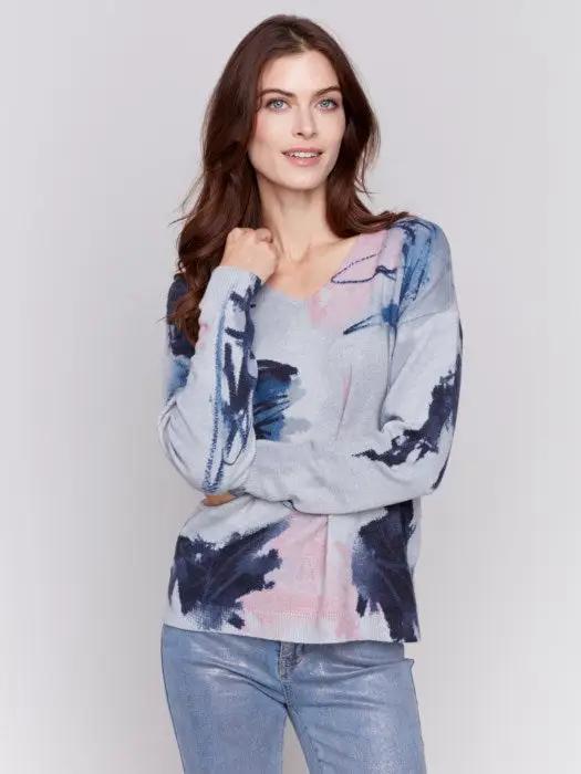 A woman with long brown hair is donning the epitome of winter comfort from Charlie B: a light grey Printed Plushy Sweater featuring an elegant watercolor-style print in pink and blue, perfectly paired with dark blue jeans.