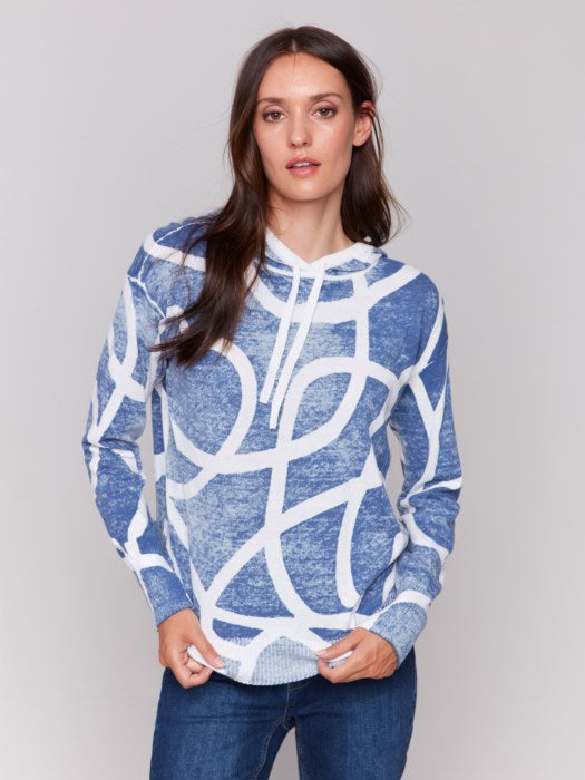 A person wearing the cozy and stylish Reverse Print Hoodie Sweater by Charlie B, featuring a blue and white pattern, pairs it with jeans against a plain gray background, exuding a playful print vibe.