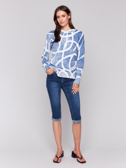 A person wearing the cozy and stylish Reverse Print Hoodie Sweater by Charlie B, featuring a blue and white pattern, pairs it with jeans against a plain gray background, exuding a playful print vibe.