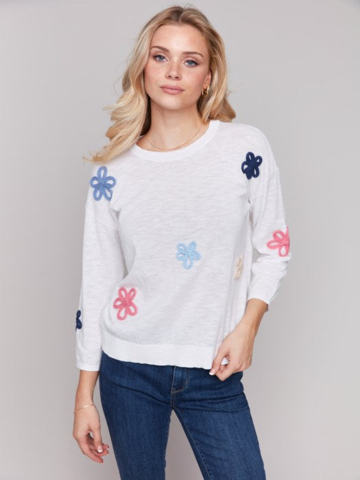 Wearing a Charlie B Cotton Sweater with Flower Patches and blue jeans, a person stands against a plain background.