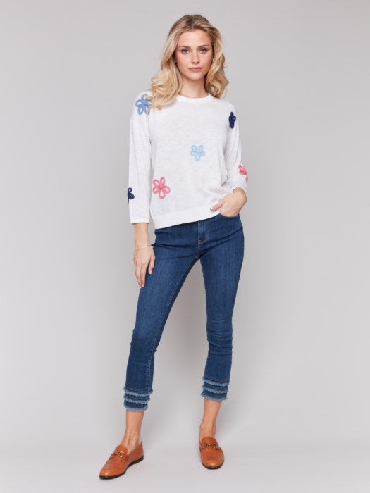 Wearing a Charlie B Cotton Sweater with Flower Patches and blue jeans, a person stands against a plain background.