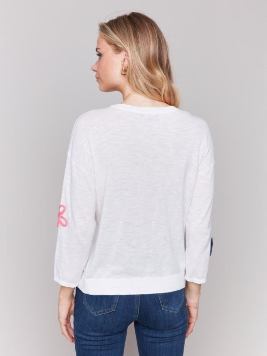 Wearing a Charlie B Cotton Sweater with Flower Patches and blue jeans, a person stands against a plain background.