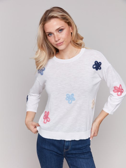 Wearing a Charlie B Cotton Sweater with Flower Patches and blue jeans, a person stands against a plain background.