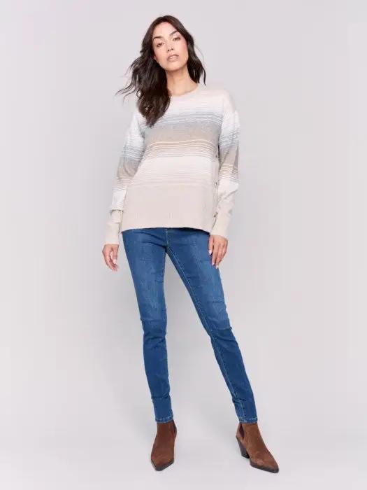 A woman wearing Charlie B's fashion-forward Crew Neck With Side Buttons, a striped long-sleeve sweater in light colors. She has long, wavy dark hair and is posing against a plain white background.