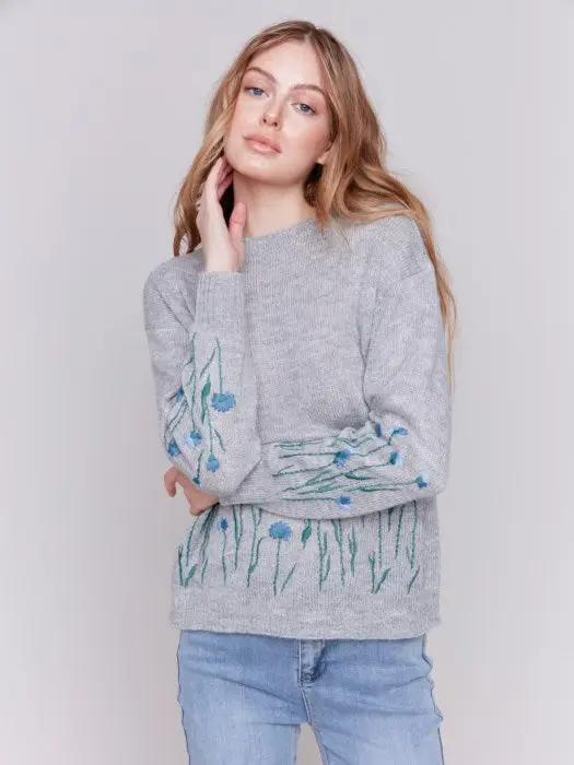 A woman with long hair is wearing the Charlie B Plush Knit Embroidered Flower Sweater featuring gray fabric adorned with blue and green floral details, paired with light blue jeans, creating a look that exudes cozy warmth.