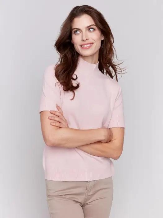 A stylish and timeless woman with long brown hair is wearing the Mock Neck Basic Top by Charlie B in light pink, featuring 3/4 sleeves, paired with beige pants. She stands with her arms crossed, smiling and looking to the side.