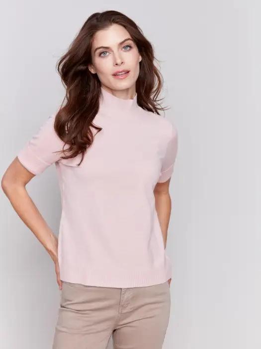 A stylish and timeless woman with long brown hair is wearing the Mock Neck Basic Top by Charlie B in light pink, featuring 3/4 sleeves, paired with beige pants. She stands with her arms crossed, smiling and looking to the side.