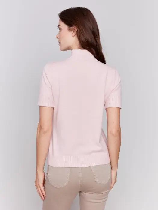 A stylish and timeless woman with long brown hair is wearing the Mock Neck Basic Top by Charlie B in light pink, featuring 3/4 sleeves, paired with beige pants. She stands with her arms crossed, smiling and looking to the side.