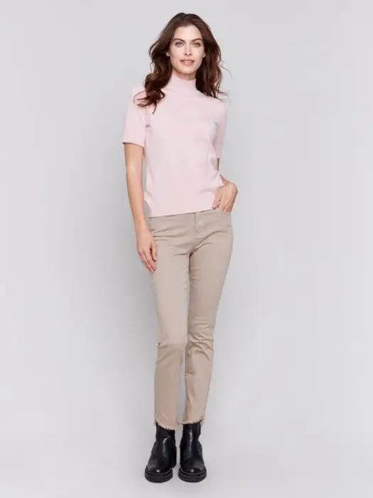 A stylish and timeless woman with long brown hair is wearing the Mock Neck Basic Top by Charlie B in light pink, featuring 3/4 sleeves, paired with beige pants. She stands with her arms crossed, smiling and looking to the side.