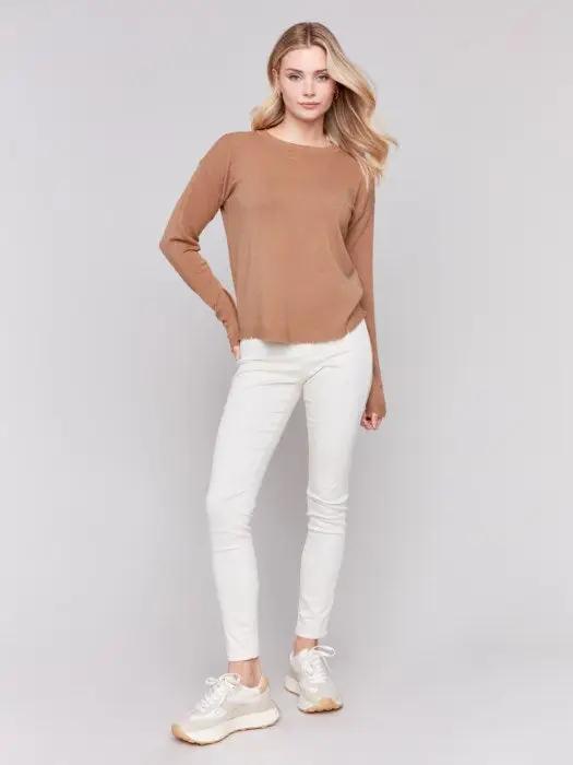 A woman wearing a stylish Round Hem Sweater by Charlie B and white pants stands against a plain background, perfect for cozy nights in.