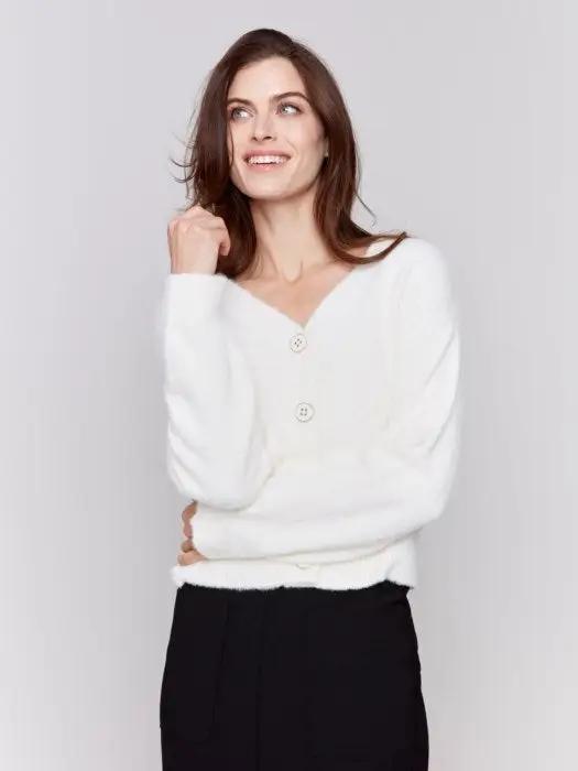 A person is styled in a cozy comfort white ensemble from Charlie B, consisting of a Short Fuzzy Knit Cardigan with buttons and paired with black pants, posed against a plain background while looking to the side with a slight smile, perfectly encapsulating this must-have wardrobe piece.