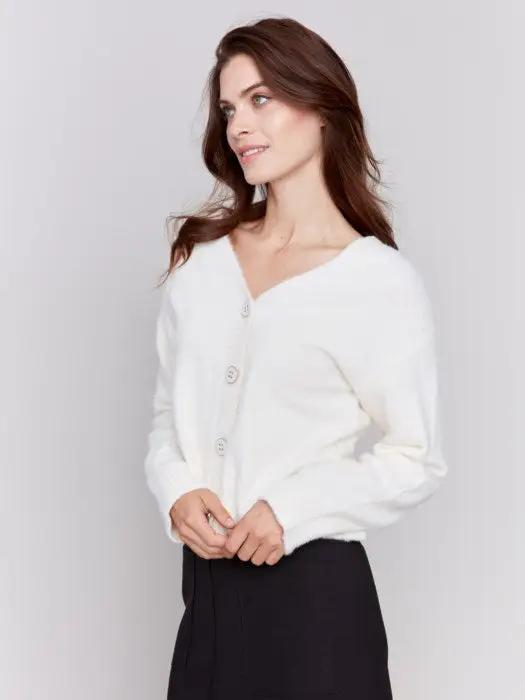 A person is styled in a cozy comfort white ensemble from Charlie B, consisting of a Short Fuzzy Knit Cardigan with buttons and paired with black pants, posed against a plain background while looking to the side with a slight smile, perfectly encapsulating this must-have wardrobe piece.