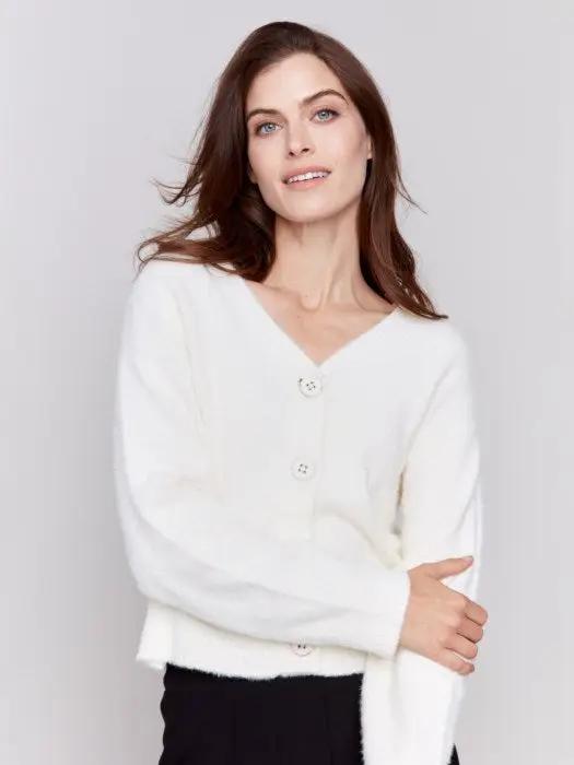 A person is styled in a cozy comfort white ensemble from Charlie B, consisting of a Short Fuzzy Knit Cardigan with buttons and paired with black pants, posed against a plain background while looking to the side with a slight smile, perfectly encapsulating this must-have wardrobe piece.