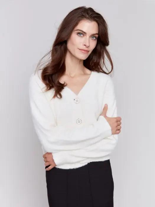 A person is styled in a cozy comfort white ensemble from Charlie B, consisting of a Short Fuzzy Knit Cardigan with buttons and paired with black pants, posed against a plain background while looking to the side with a slight smile, perfectly encapsulating this must-have wardrobe piece.