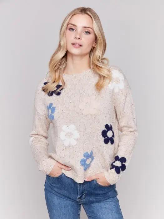 A person with long blonde hair wearing a Charlie B Neps Yarn Knit Sweater adorned with colorful flower patterns and blue jeans stands with hands in pockets against a plain background, perfect for cooler weather.