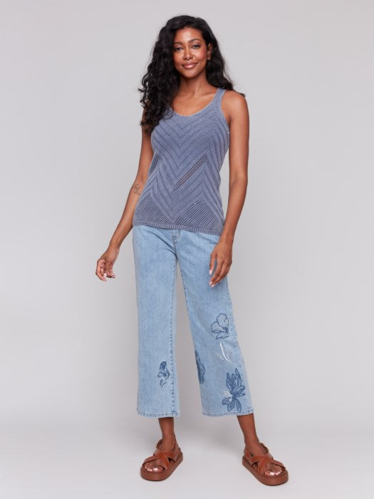 A person smiles, dressed in a chic Charlie B Chevron Pointelle Knit Camisole and light blue jeans, set against a gray background.