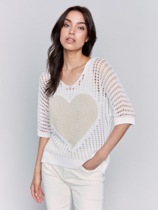 A woman poses against a plain backdrop, wearing the Fishnet V-Neck Sweater with Gold Heart by Charlie B, stylishly paired with light pants.