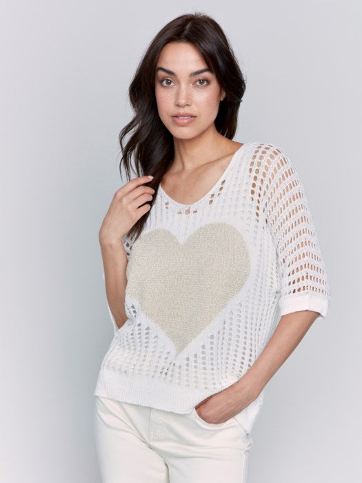 A woman poses against a plain backdrop, wearing the Fishnet V-Neck Sweater with Gold Heart by Charlie B, stylishly paired with light pants.