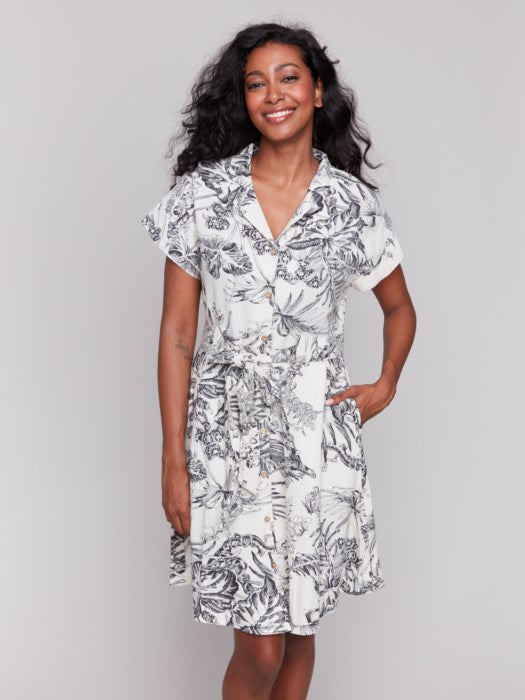 A person models the Charlie B Linen Blend Short Sleeve Button-Front Dress, featuring a black and white floral pattern that reaches knee-length. They smile with hands in pockets, set against a plain background.