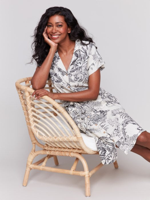 A person models the Charlie B Linen Blend Short Sleeve Button-Front Dress, featuring a black and white floral pattern that reaches knee-length. They smile with hands in pockets, set against a plain background.