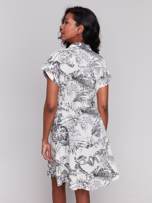 A person models the Charlie B Linen Blend Short Sleeve Button-Front Dress, featuring a black and white floral pattern that reaches knee-length. They smile with hands in pockets, set against a plain background.