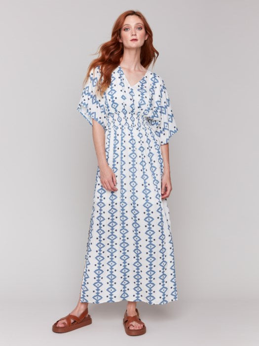 A fashionista in a Charlie B Printed Dolman Sleeve Maxi Dress with Side Slits stands sideways, eyes on the camera, hand on hip, paired with chic white heeled sandals.