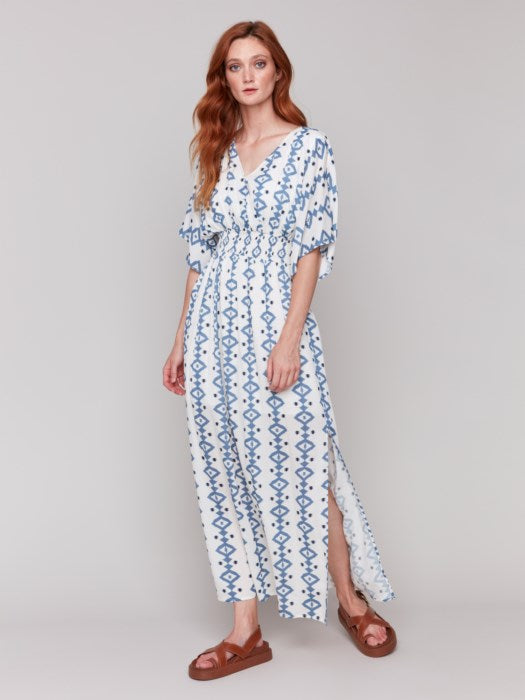 A fashionista in a Charlie B Printed Dolman Sleeve Maxi Dress with Side Slits stands sideways, eyes on the camera, hand on hip, paired with chic white heeled sandals.