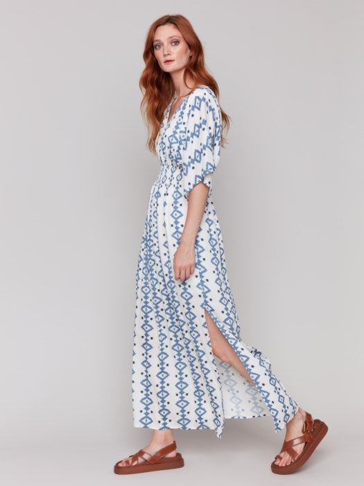 A fashionista in a Charlie B Printed Dolman Sleeve Maxi Dress with Side Slits stands sideways, eyes on the camera, hand on hip, paired with chic white heeled sandals.