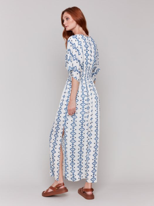 A fashionista in a Charlie B Printed Dolman Sleeve Maxi Dress with Side Slits stands sideways, eyes on the camera, hand on hip, paired with chic white heeled sandals.