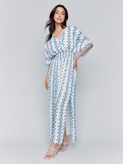 A fashionista in a Charlie B Printed Dolman Sleeve Maxi Dress with Side Slits stands sideways, eyes on the camera, hand on hip, paired with chic white heeled sandals.