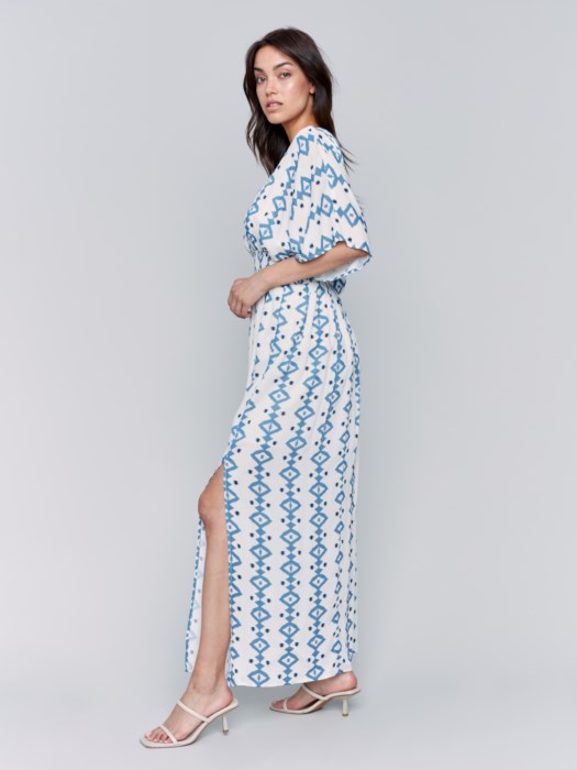 A fashionista in a Charlie B Printed Dolman Sleeve Maxi Dress with Side Slits stands sideways, eyes on the camera, hand on hip, paired with chic white heeled sandals.