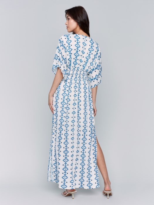 A fashionista in a Charlie B Printed Dolman Sleeve Maxi Dress with Side Slits stands sideways, eyes on the camera, hand on hip, paired with chic white heeled sandals.