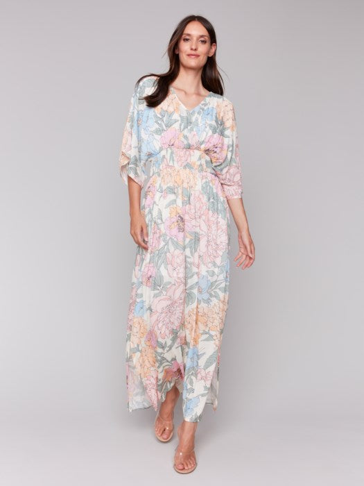 A woman stands against a neutral backdrop wearing the Dolman Sleeve Maxi Dress with Side Slits by Charlie B, paired with sandals. The dress features a flowing, pastel-toned floral design.