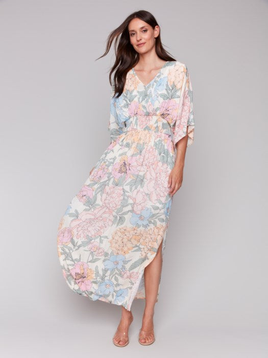 A woman stands against a neutral backdrop wearing the Dolman Sleeve Maxi Dress with Side Slits by Charlie B, paired with sandals. The dress features a flowing, pastel-toned floral design.