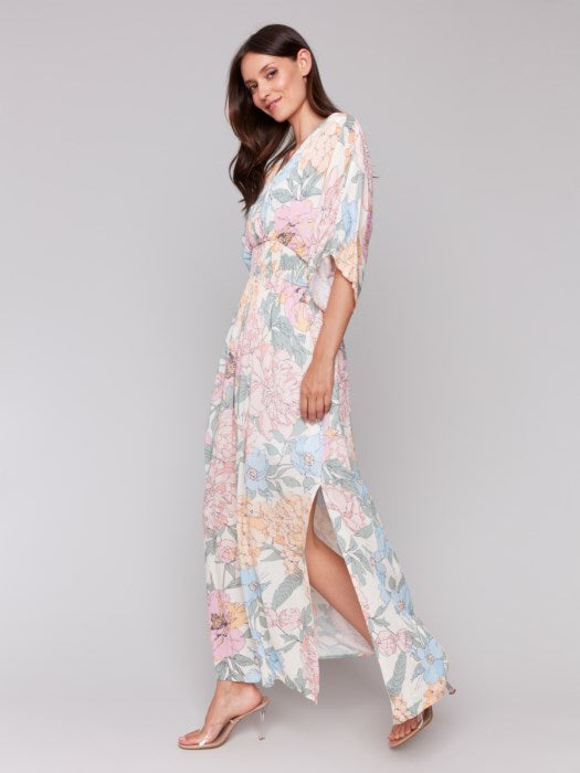 A woman stands against a neutral backdrop wearing the Dolman Sleeve Maxi Dress with Side Slits by Charlie B, paired with sandals. The dress features a flowing, pastel-toned floral design.