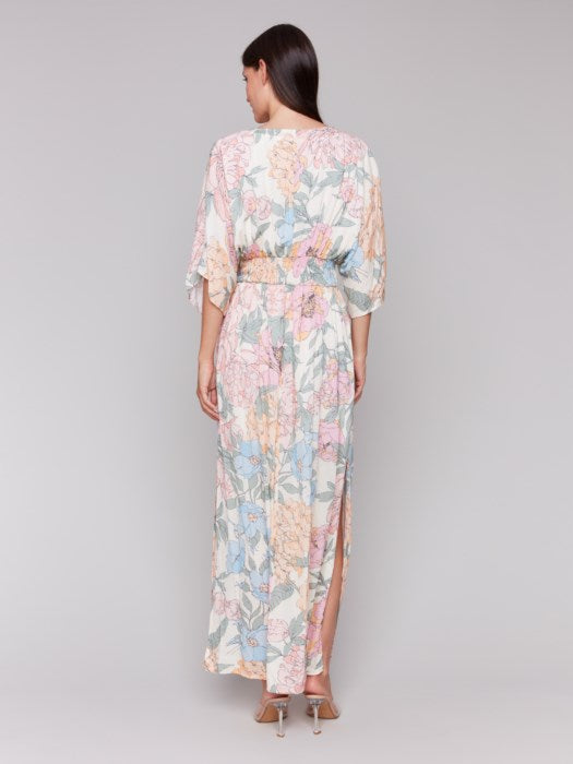 A woman stands against a neutral backdrop wearing the Dolman Sleeve Maxi Dress with Side Slits by Charlie B, paired with sandals. The dress features a flowing, pastel-toned floral design.