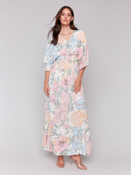 A woman stands against a neutral backdrop wearing the Dolman Sleeve Maxi Dress with Side Slits by Charlie B, paired with sandals. The dress features a flowing, pastel-toned floral design.