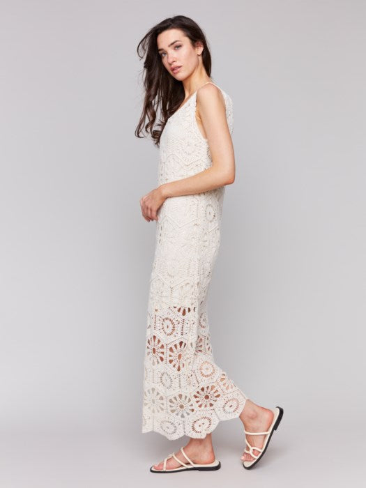 A woman models the Charlie B Cotton Crochet Sleeveless Maxi Dress, showcasing its feminine charm in a long white design. She stands sideways with hands by her sides, wearing black sandals against a plain grey backdrop.