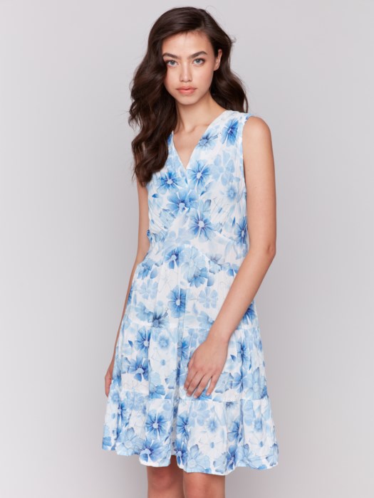 A woman in a Charlie B Printed Sleeveless V-Neck Tiered Dress stands against a plain background. The light blue and white floral design offers an effortlessly chic look, paired with nude heels, her hair cascading elegantly over her shoulders.