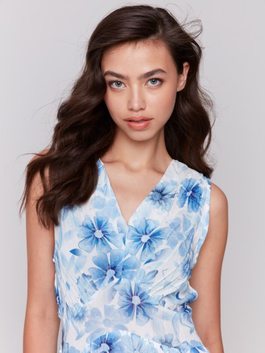 A woman in a Charlie B Printed Sleeveless V-Neck Tiered Dress stands against a plain background. The light blue and white floral design offers an effortlessly chic look, paired with nude heels, her hair cascading elegantly over her shoulders.