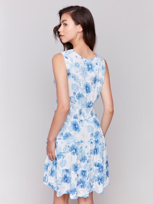 A woman in a Charlie B Printed Sleeveless V-Neck Tiered Dress stands against a plain background. The light blue and white floral design offers an effortlessly chic look, paired with nude heels, her hair cascading elegantly over her shoulders.