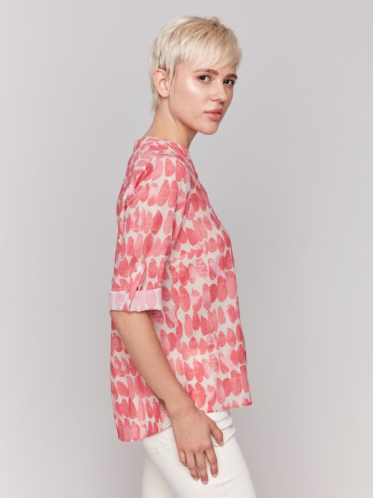 A person with short blonde hair, wearing a Charlie B printed viscose half-button blouse in pink and white and white pants, stands against a plain background.