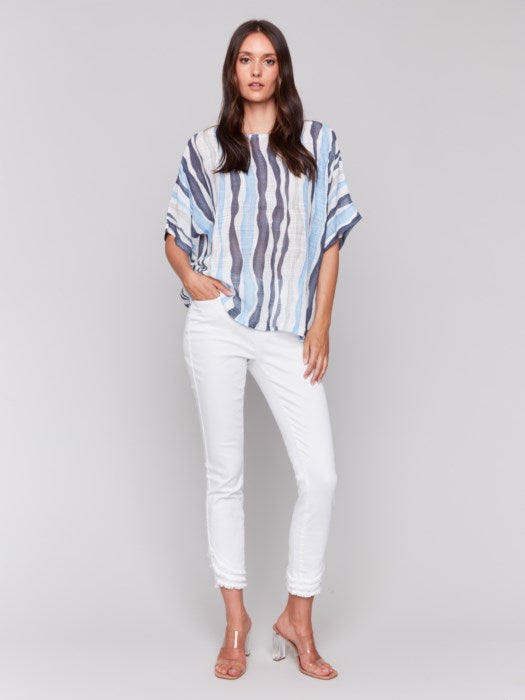 A person models Charlie B's Cotton Gauze Dolman Top in blue and white, paired with white pants against a gray background, capturing the summer vibe with its airy fabric.