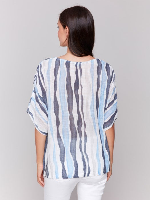 A person models Charlie B's Cotton Gauze Dolman Top in blue and white, paired with white pants against a gray background, capturing the summer vibe with its airy fabric.