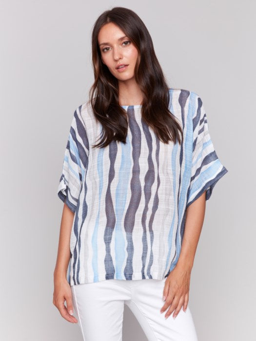 A person models Charlie B's Cotton Gauze Dolman Top in blue and white, paired with white pants against a gray background, capturing the summer vibe with its airy fabric.