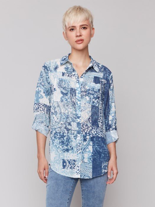 A person with short blond hair is wearing a Charlie B Printed Crinkle Button-Front Blouse in blue and white, paired with jeans, standing against a plain background.