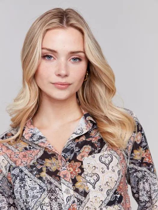 A woman with blonde hair is seated on the floor with one knee up, wearing a Front Button Up Paisley Blouse by Charlie B, along with blue jeans and black boots. She is looking straight at the camera against a plain background.